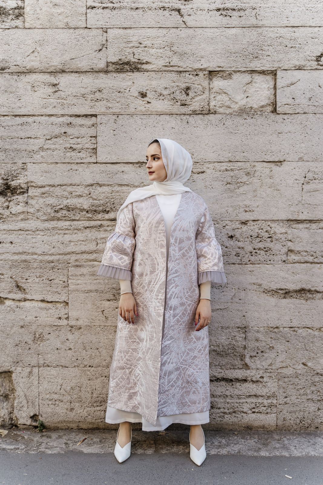 Woman in a pale patterned coat standing against a stone wall